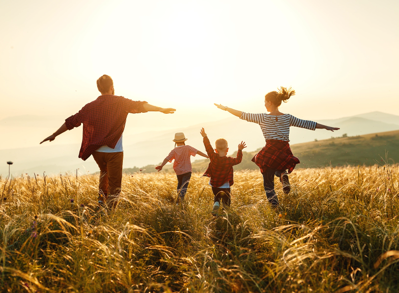 A family of with two young children running carefree through a field with their arms out .
